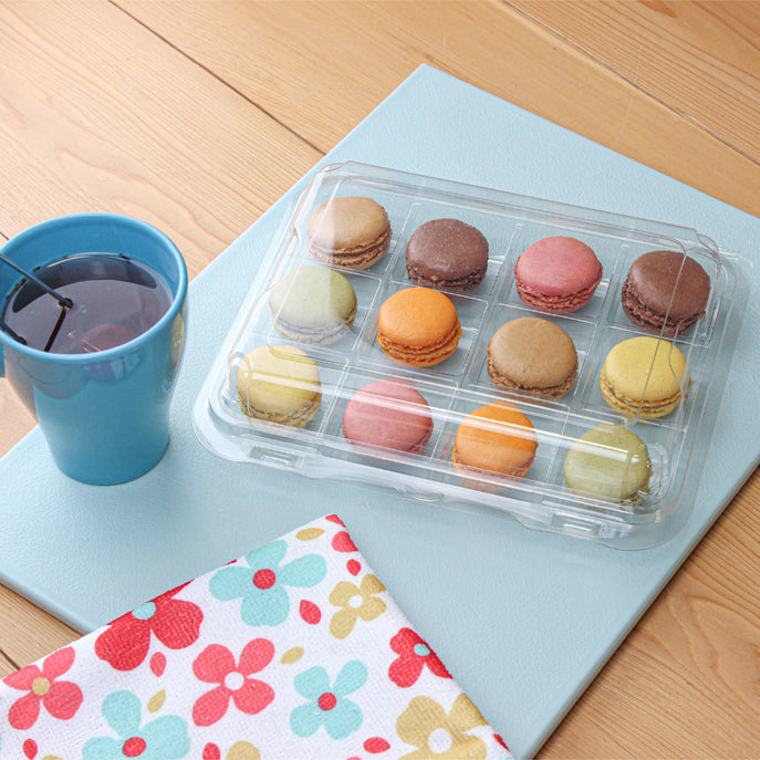 12 pack of Macarons in a container next to a flowered naplin and a blue cup with tea