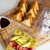 square food packaging made of clear, compostable plastic containing deli goods, fresh fruit, and baked goods